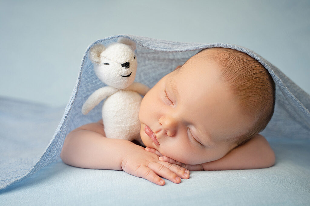baby on blue background under blanket with white teddy bear